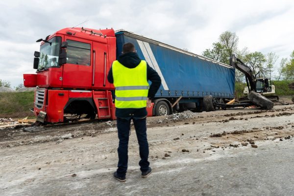 A man in a reflective vest observes a damaged red semi-truck and trailer after a highway accident cleanup