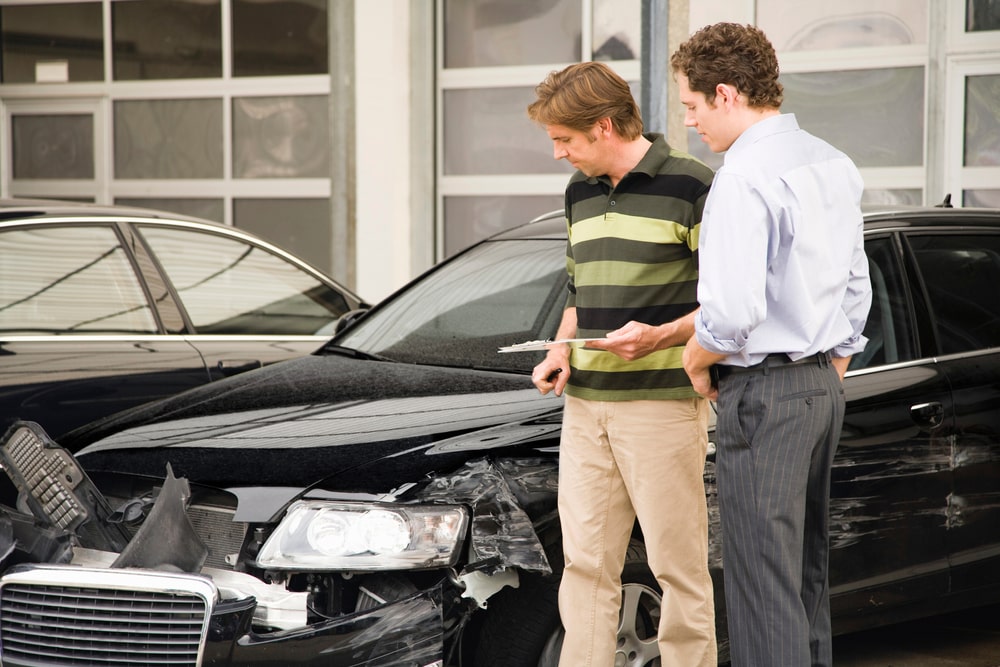Two men are reviewing damage on a black car after an accident outside a repair shop