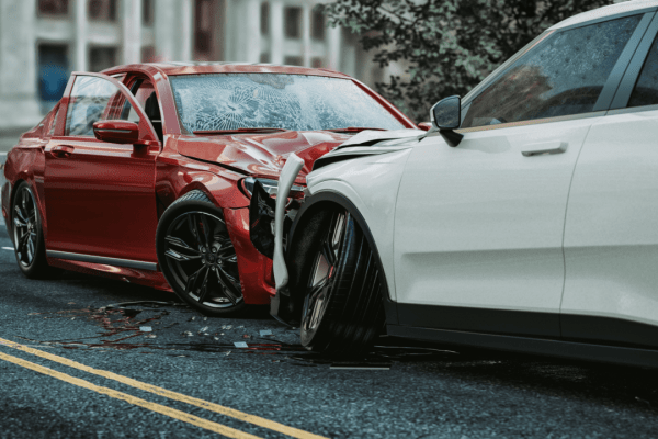 Two damaged cars after a collision on a city street - a red sedan with front-end damage and cracked windshield, and a white SUV with side damage, with debris scattered on the road