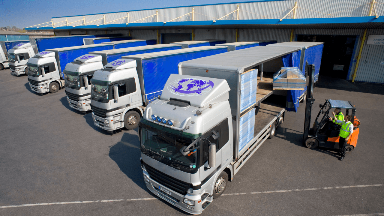 Fleet of white delivery trucks parked at a distribution center loading dock with workers operating a forklift nearby