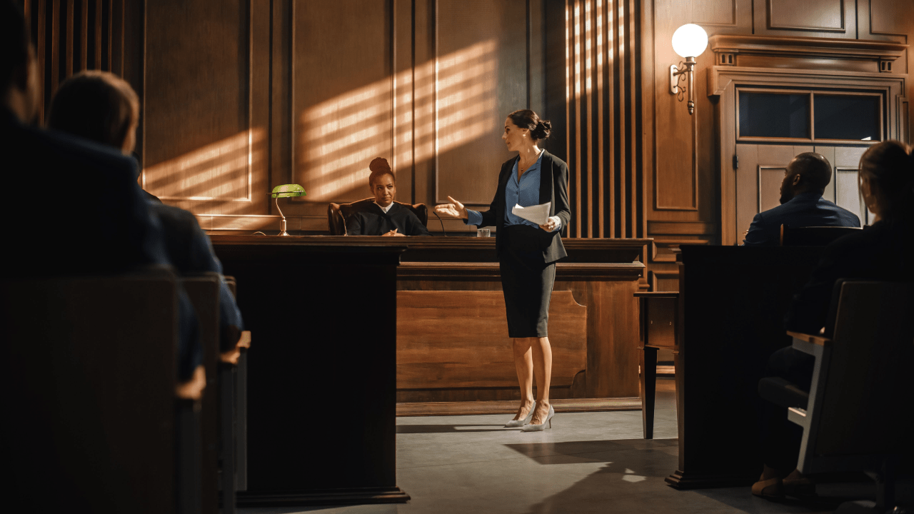 Female attorney in professional attire presenting case to judge in formal courtroom with gallery of observers