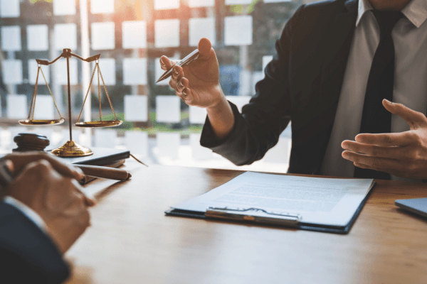 Professional in dark suit consulting with client over legal documents at wooden desk, with scales of justice and gavel visible in law office setting