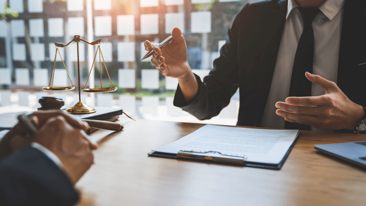Professional in dark suit consulting with client over legal documents at wooden desk, with scales of justice and gavel visible in law office setting