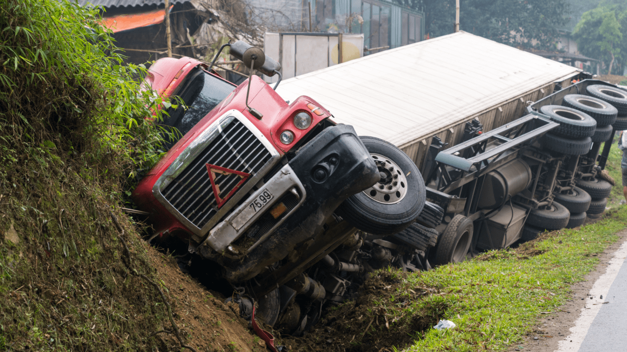 Overturned truck accident with red cab and white trailer lying on its side beside the road in a rural area