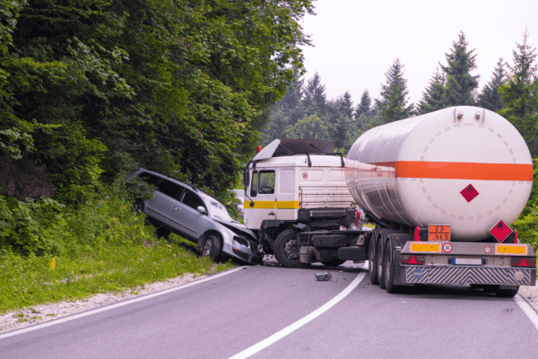 Traffic accident between a silver car and white tanker truck with hazmat placards on a forest road