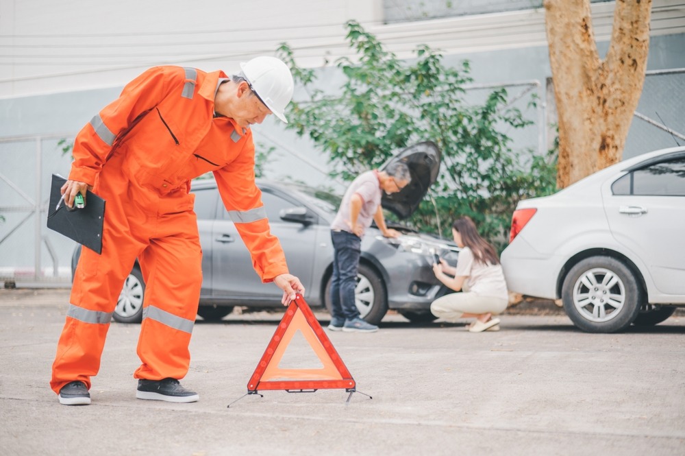 Accident scene with safety inspector placing a warning triangle as two individuals inspect vehicle damage.