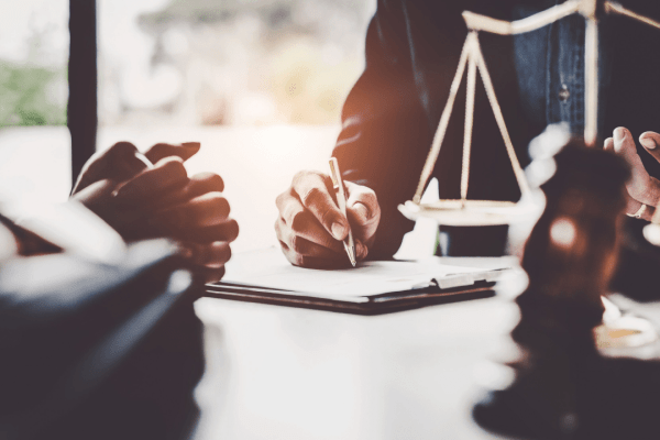 Person signing legal documents at desk with scales of justice visible in law office setting