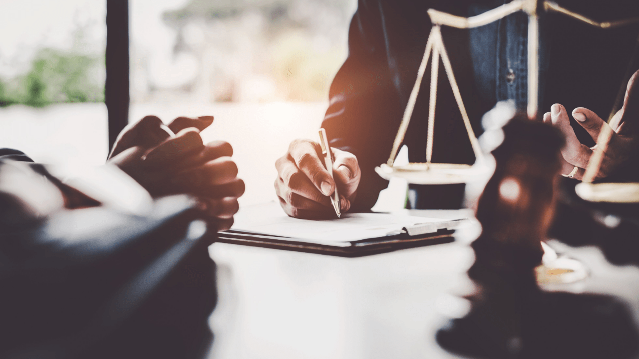 Person signing legal documents at desk with scales of justice visible in law office setting