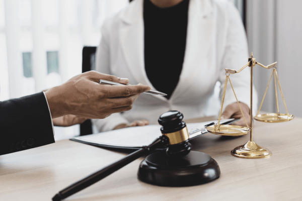 Person in a suit showing a phone to a medical professional in a white coat at a desk with legal items, including a gavel and scales of justice