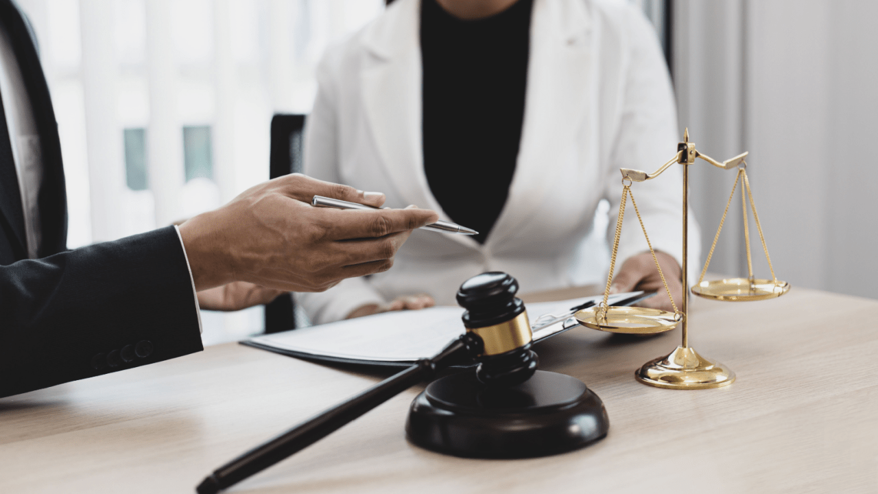 Person in a suit showing a phone to a medical professional in a white coat at a desk with legal items, including a gavel and scales of justice