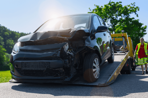 Damaged black car with front-end collision damage being loaded onto a yellow tow truck by an operator in a safety vest