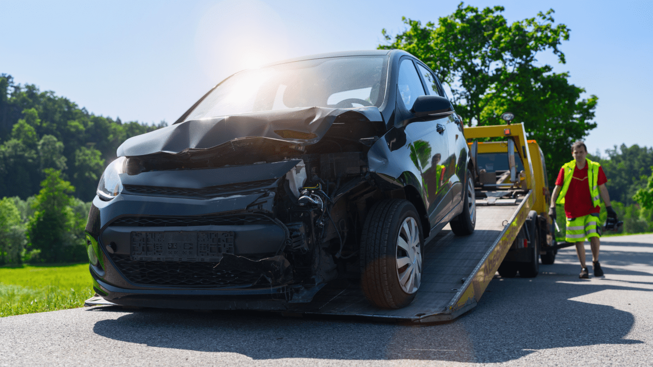 Damaged black car with front-end collision damage being loaded onto a yellow tow truck by an operator in a safety vest