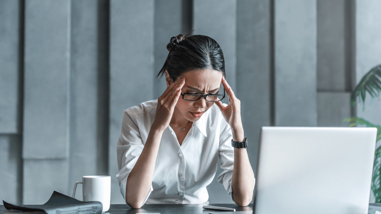 Professional woman in white shirt and glasses holding her temples with both hands while sitting at office desk with laptop, appearing to have a headache or experiencing work stress