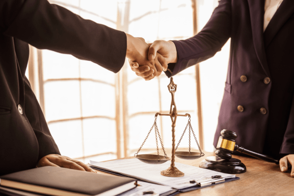 Two lawyers shaking hands over insurance and settlement factors agreement with scales of justice and gavel on desk