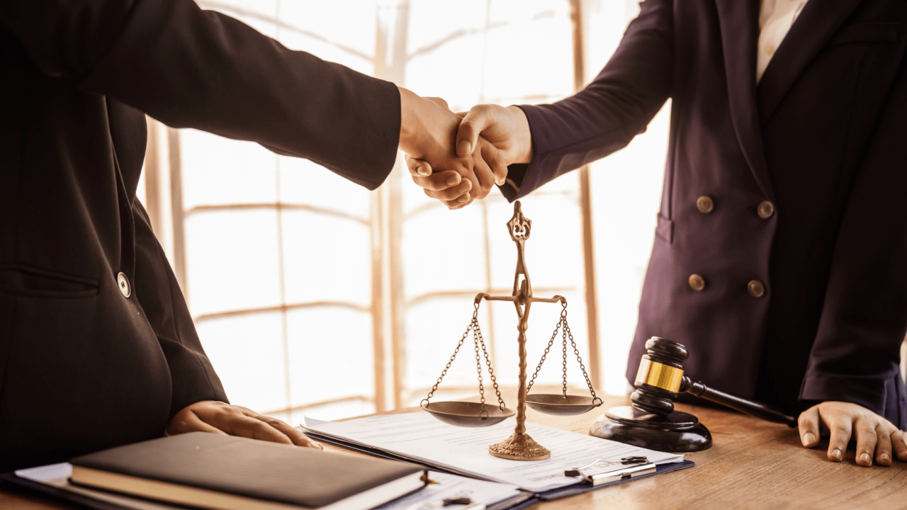 Two lawyers shaking hands over insurance and settlement factors agreement with scales of justice and gavel on desk