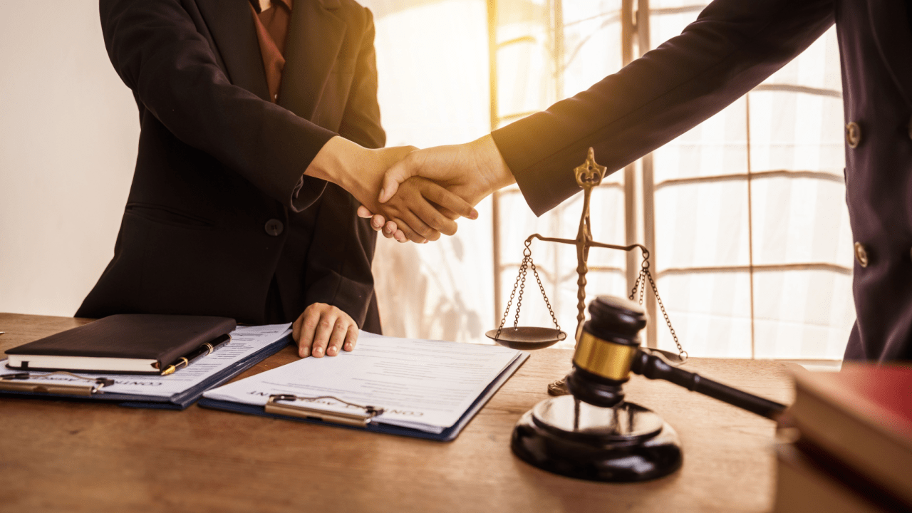 Two attorneys shaking hands over legal documents discussing what is a reasonable settlement offer with scales of justice and gavel on desk